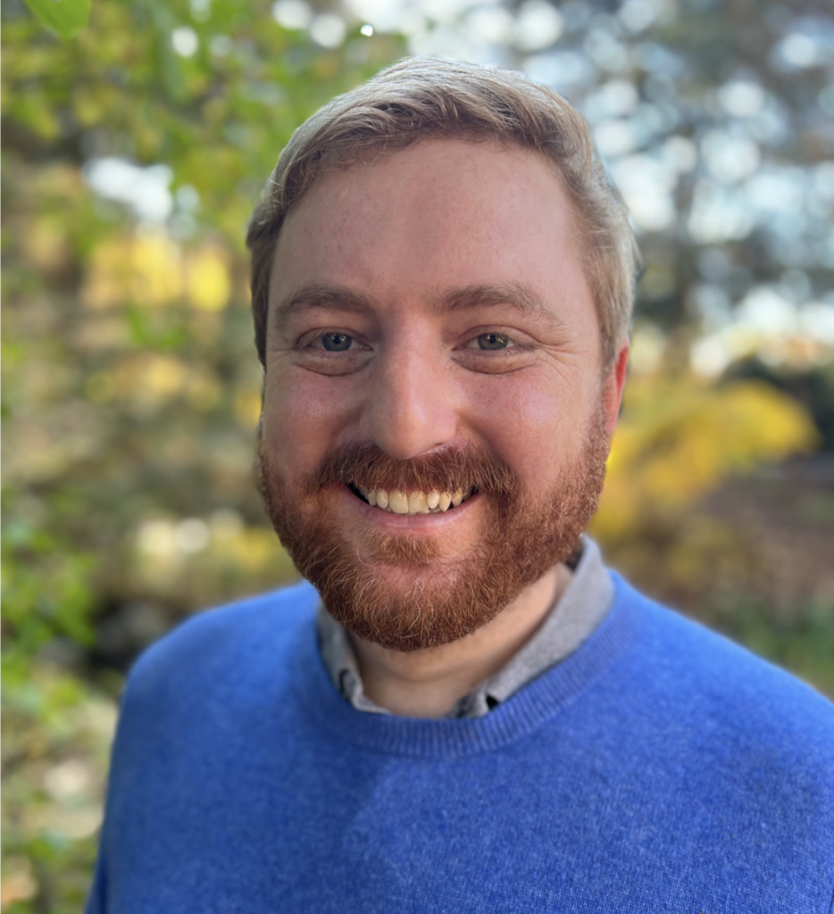 Man with redish blond hair and a beard wearing a blue sweater is smiling.