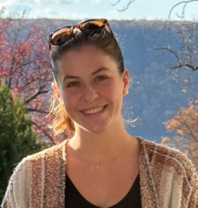 Young woman with light brown hair smiling and standing in front of a scenic background.