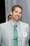Man with short brown hair wearing a light colored suit and green tie.